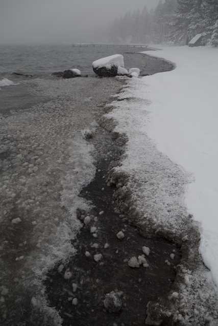 A snowy, rocky shoreline with icy water and a foggy background.