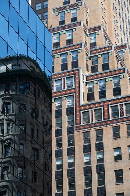 A tan brick building with numerous rectangular windows stands adjacent to a glass-faced building reflecting other structures.