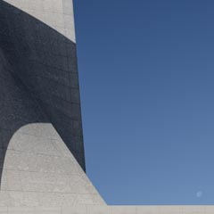 A tall stone building with a curved shadow on its side under a clear blue sky with the moon visible above.