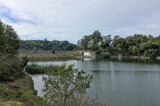 An earthen dam spans across a lake, with a concrete spillway and a metal railing along its edge.