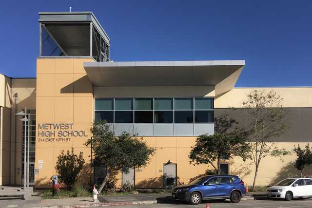 A large building with a flat roof and many windows, labeled "MetWest High School", under a clear blue sky.
