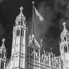 A flag flies atop a Gothic-style building with ornate spires and intricate stonework, set against a cloudy sky.