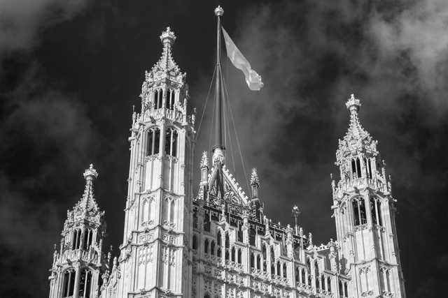 A flag flies atop a Gothic-style building with ornate spires and intricate stonework, set against a cloudy sky.