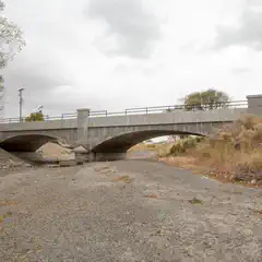 A concrete bridge over a dry riverbed with sparse vegetation and cloudy sky.