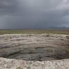 A large, circular rock formation with layered walls overlooks a flat, grassy plain under a heavy, overcast sky.