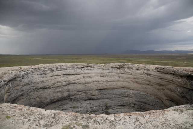 A large, circular rock formation with layered walls overlooks a flat, grassy plain under a heavy, overcast sky.