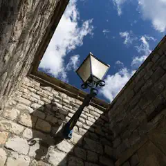 A black lamppost with a glass lantern is mounted on a stone wall, framed by weathered masonry under a partly cloudy sky.