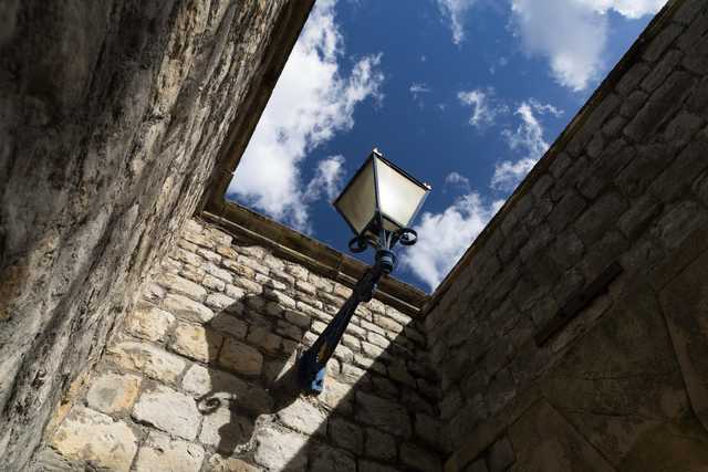 A black lamppost with a glass lantern is mounted on a stone wall, framed by weathered masonry under a partly cloudy sky.