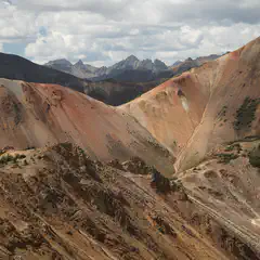 A rugged mountain pass with steep, multicolored rock formations and sparse vegetation under a partly cloudy sky.