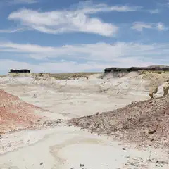 A dry, eroded valley with layered rock formations, reddish slopes, and scattered boulders under a partly cloudy sky.