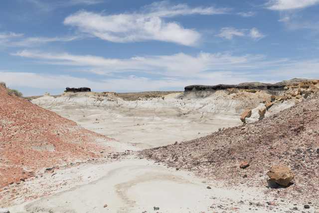A dry, eroded valley with layered rock formations, reddish slopes, and scattered boulders under a partly cloudy sky.