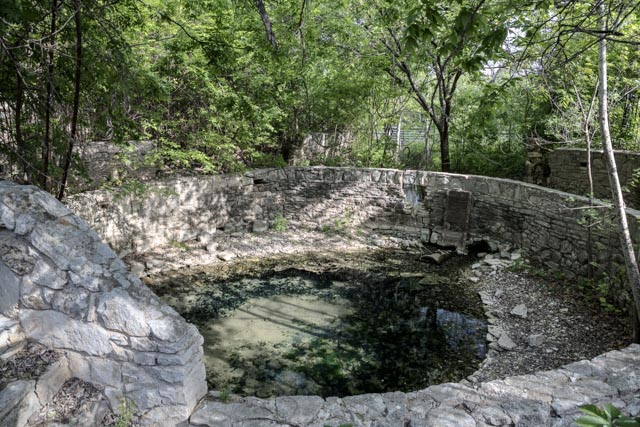 An old, circular stone structure enclosing a shallow pool of water with algae and surrounded by trees.