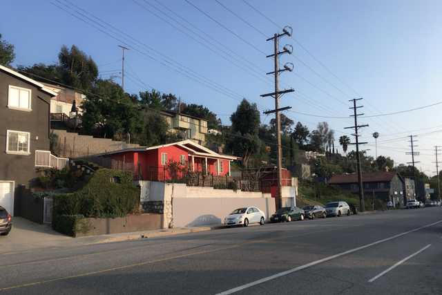 An urban street is lined with houses and cars, set against a backdrop of trees and power lines.