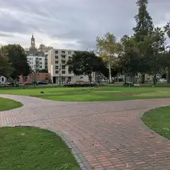 A brick walkway curves through a grassy park with trees and buildings in the background.