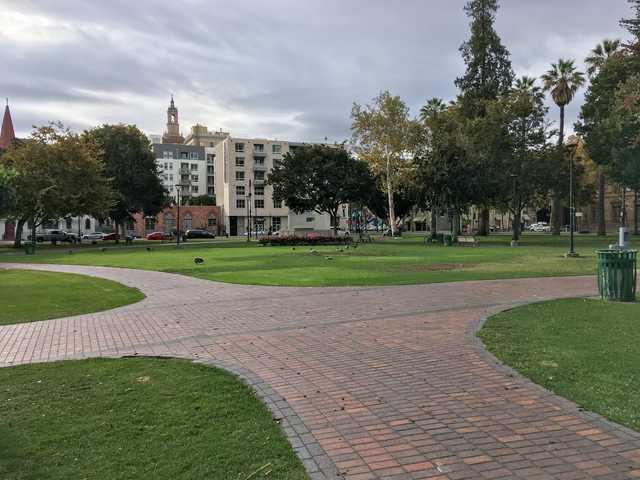 A brick walkway curves through a grassy park with trees and buildings in the background.