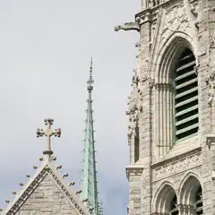 A stone building features pointed arches, a cross-topped spire, and green metal accents against a blue sky.
