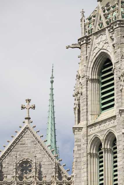 A stone building features pointed arches, a cross-topped spire, and green metal accents against a blue sky.