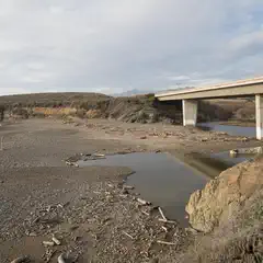 A concrete bridge crosses a narrow waterway leading to a gravelly beach littered with driftwood, with low hills in the background.