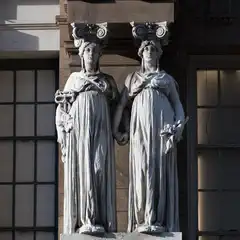 Two stone caryatids, each supporting an Ionic capital, stand side by side on a building facade, flanked by multi-paned windows.
