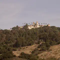 A grand, multi-towered estate crowns a hilltop, surrounded by dense greenery and open fields, under a clear sky.