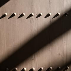 A wooden panel with evenly spaced, small round protrusions, illuminated by shadows cast by slats above it.