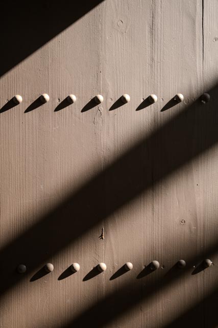 A wooden panel with evenly spaced, small round protrusions, illuminated by shadows cast by slats above it.