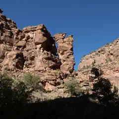 A towering red rock formation rises against a clear blue sky, with trees dotting the rugged landscape below.