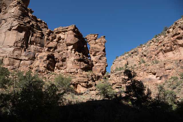 A towering red rock formation rises against a clear blue sky, with trees dotting the rugged landscape below.