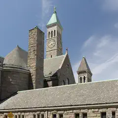 A stone church with a prominent clock tower and steeply pitched roofs stands against a clear blue sky.