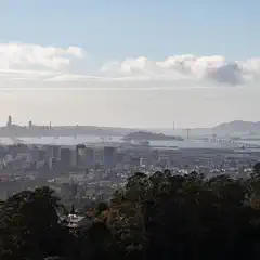 A large suspension bridge spans the bay between a small island and a city skyline with numerous tall buildings.