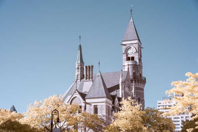 A stone building with a prominent clock tower and multiple spires rises above golden-leaved trees under a clear blue sky.