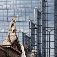 A stone statue of a figure holding a trident stands atop a classical building, with a modern glass skyscraper in the background.
