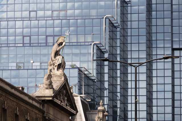 A stone statue of a figure holding a trident stands atop a classical building, with a modern glass skyscraper in the background.