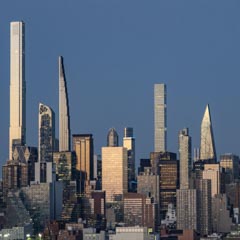A dense urban skyline featuring a mix of modern skyscrapers and illuminated buildings under a clear blue sky.