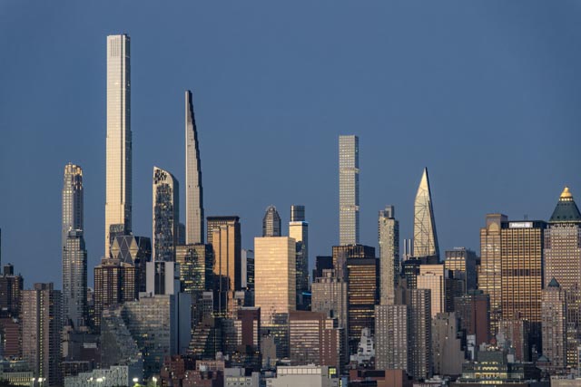 A dense urban skyline featuring a mix of modern skyscrapers and illuminated buildings under a clear blue sky.
