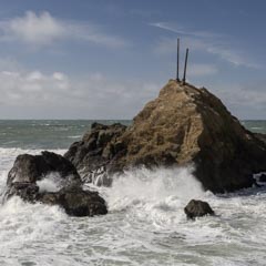 A rocky outcrop rises from turbulent ocean waves under a partly cloudy sky. Two poles extend from the top of the rock.