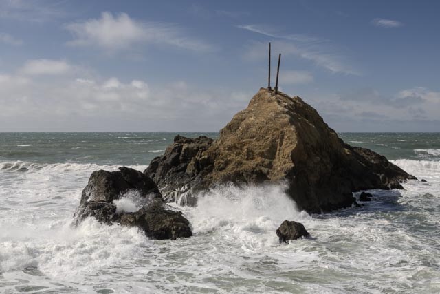 A rocky outcrop rises from turbulent ocean waves under a partly cloudy sky. Two poles extend from the top of the rock.