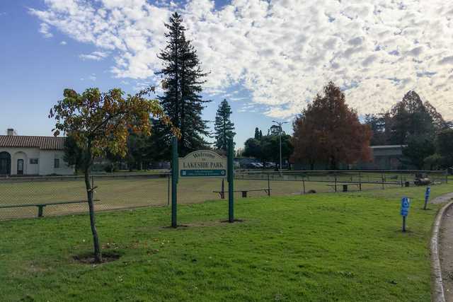 A sign for Lakeside Park stands on a grassy area with trees and a building in the background under a partly cloudy sky.