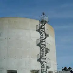 A metal spiral staircase with a railing is attached to the side of a large, cylindrical concrete structure.