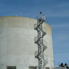 A metal spiral staircase with a railing is attached to the side of a large, cylindrical concrete structure.