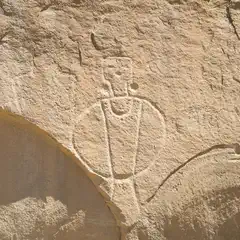 A petroglyph carved into light-colored rock depicts a stylized human figure with a rounded body, vertical lines, and a small head with circular eyes.