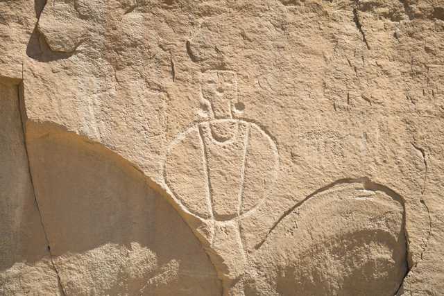 A petroglyph carved into light-colored rock depicts a stylized human figure with a rounded body, vertical lines, and a small head with circular eyes.