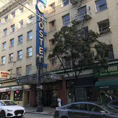 An ornate blue sign with gold lettering reads 'HOSTEL' and features a logo at the top, set against a tan brick building with numerous windows.