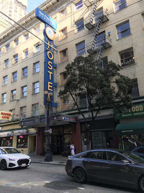An ornate blue sign with gold lettering reads 'HOSTEL' and features a logo at the top, set against a tan brick building with numerous windows.