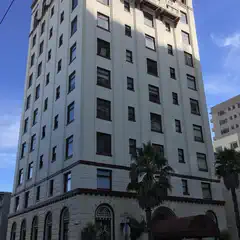 A tall, white building sits atop a street corner, featuring numerous windows, palm trees, parked cars, and a sidewalk.