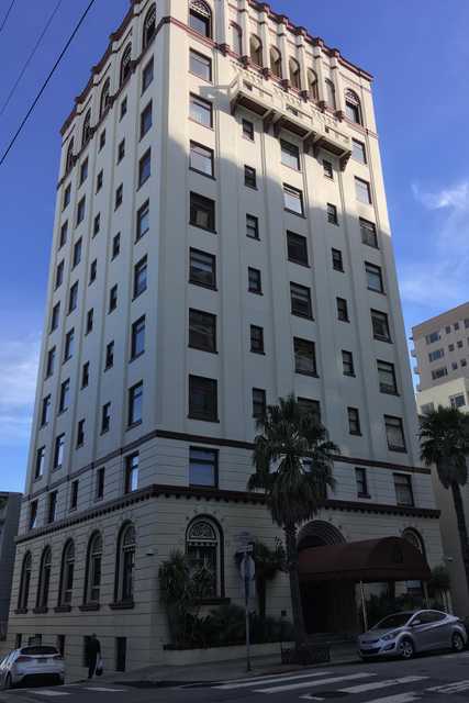 A tall, white building sits atop a street corner, featuring numerous windows, palm trees, parked cars, and a sidewalk.
