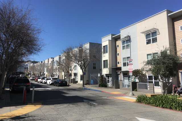 A row of three-story buildings lines a street with parked cars and bare trees.