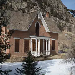 A red-brick house with white trim and a brown roof sits on a hillside, surrounded by trees and snow-covered ground.