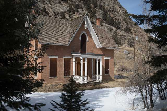 A red-brick house with white trim and a brown roof sits on a hillside, surrounded by trees and snow-covered ground.