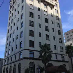A tall, white building sits atop a street corner, featuring numerous windows, palm trees, parked cars, and a sidewalk.